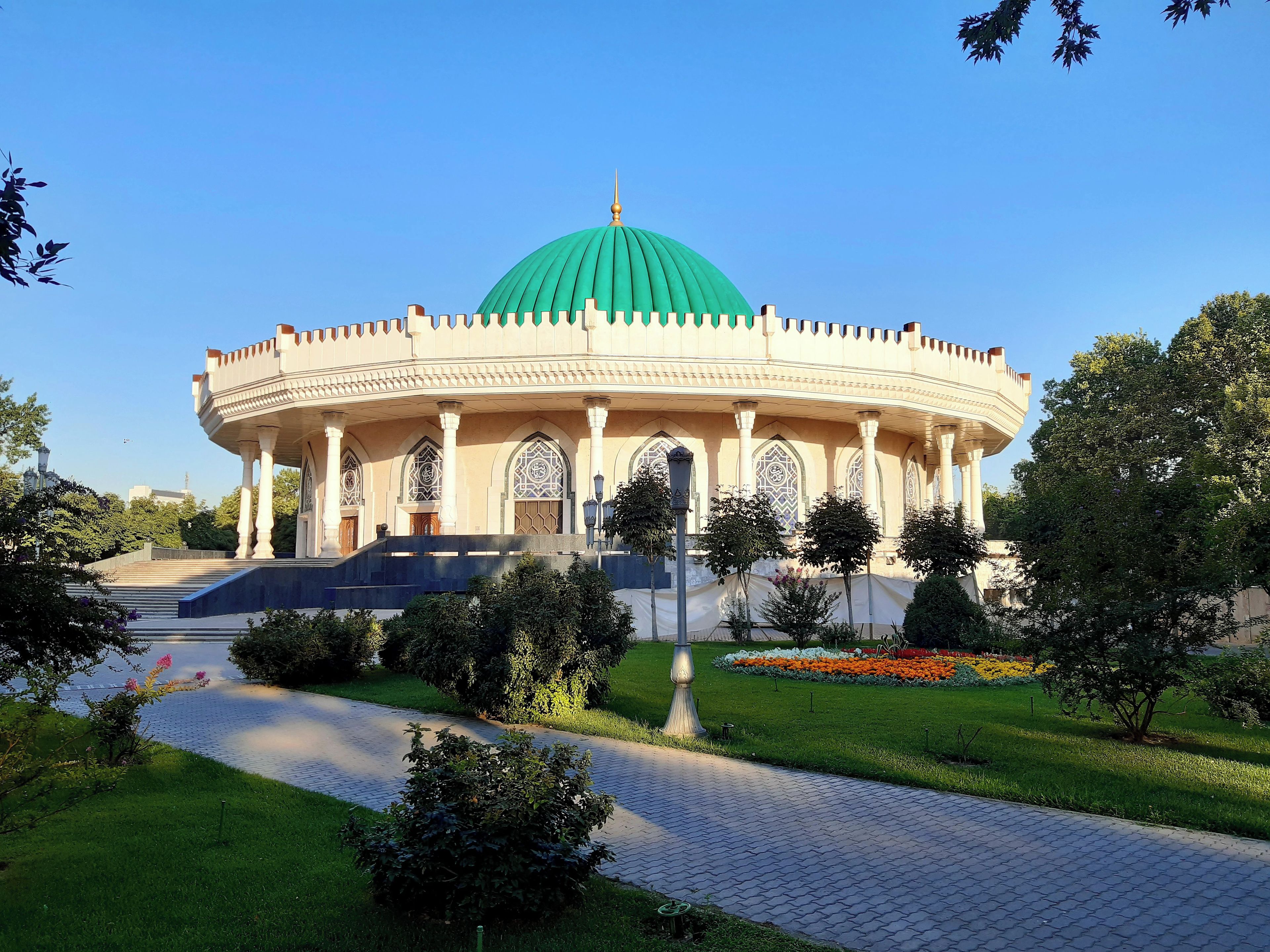 A large, circular building with a green dome and multiple arched windows, surrounded by a landscaped garden with a paved walkway.