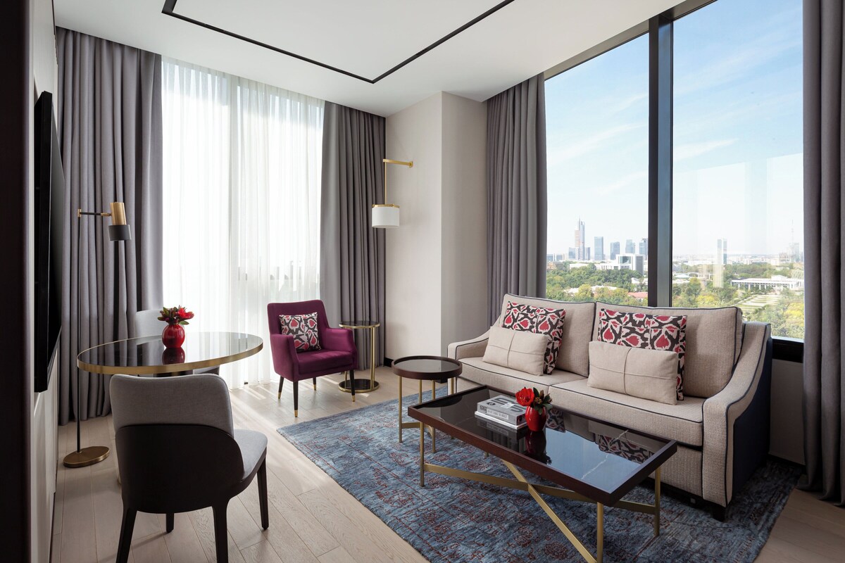 A modern living room with a sofa, armchair, coffee table, and a view of the cityscape through large windows.