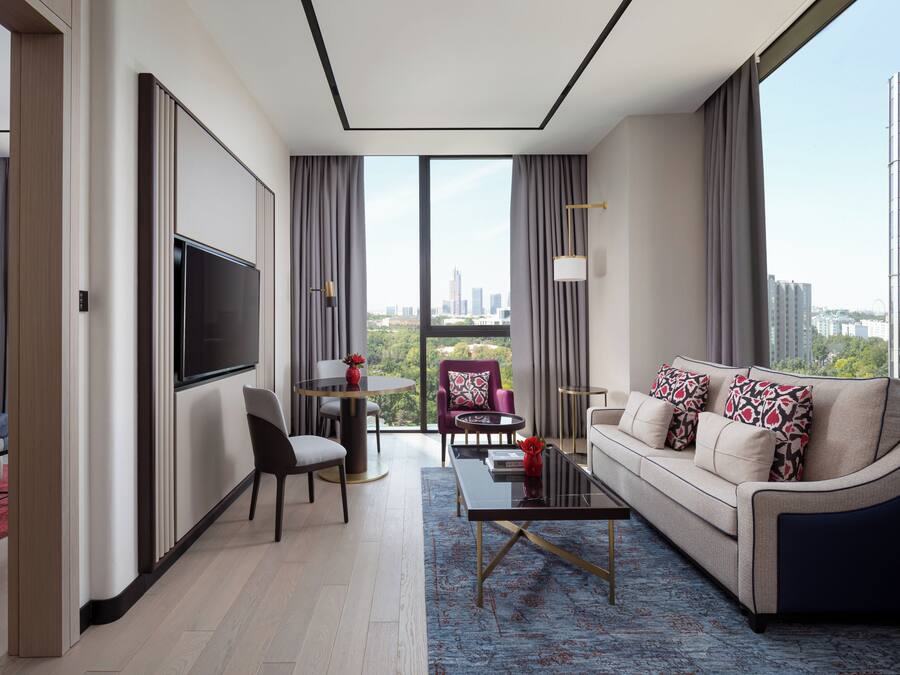 A modern hotel room with a bed, a sofa, a TV, a dining area, and a view of the cityscape.