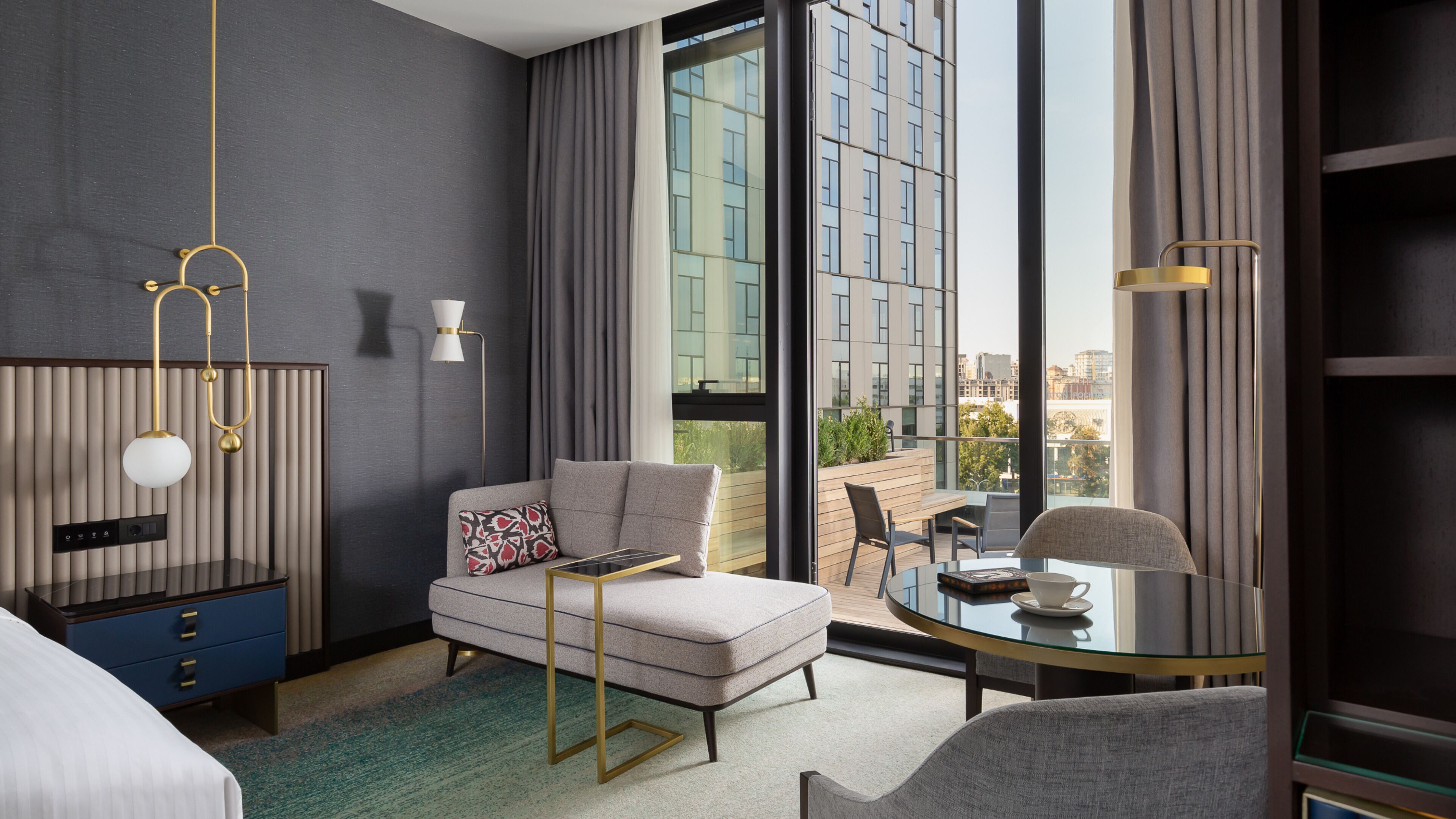 Une chambre d’hôtel moderne équipée d’un canapé, d’une table en verre, d’un lit, d’un bureau et offrant une vue sur la ville grâce à de grandes fenêtres.