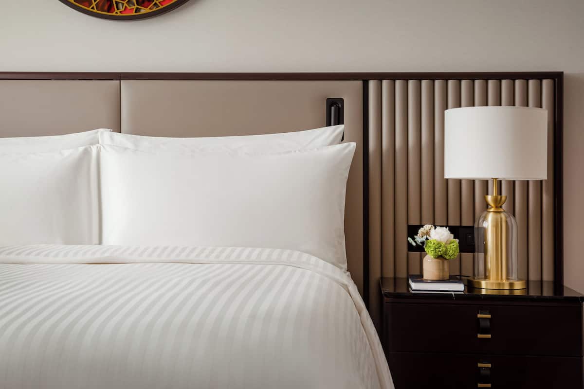 A neatly made bed with white linens, a bedside table with a lamp, and a decorative vase with flowers.