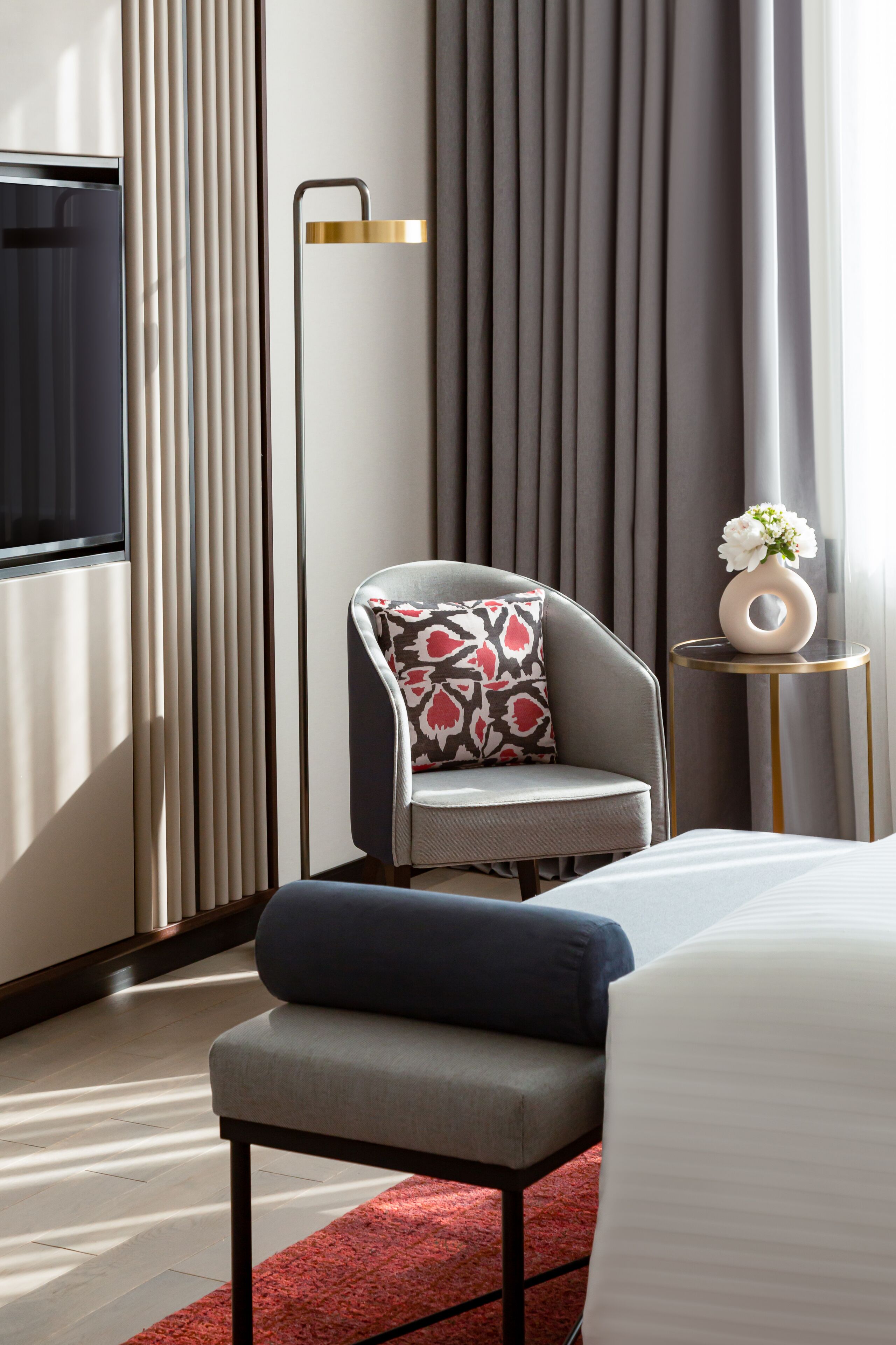 A modern hotel room with a large bed, a grey armchair with a patterned pillow, a small side table with a vase of flowers, and a floor lamp.