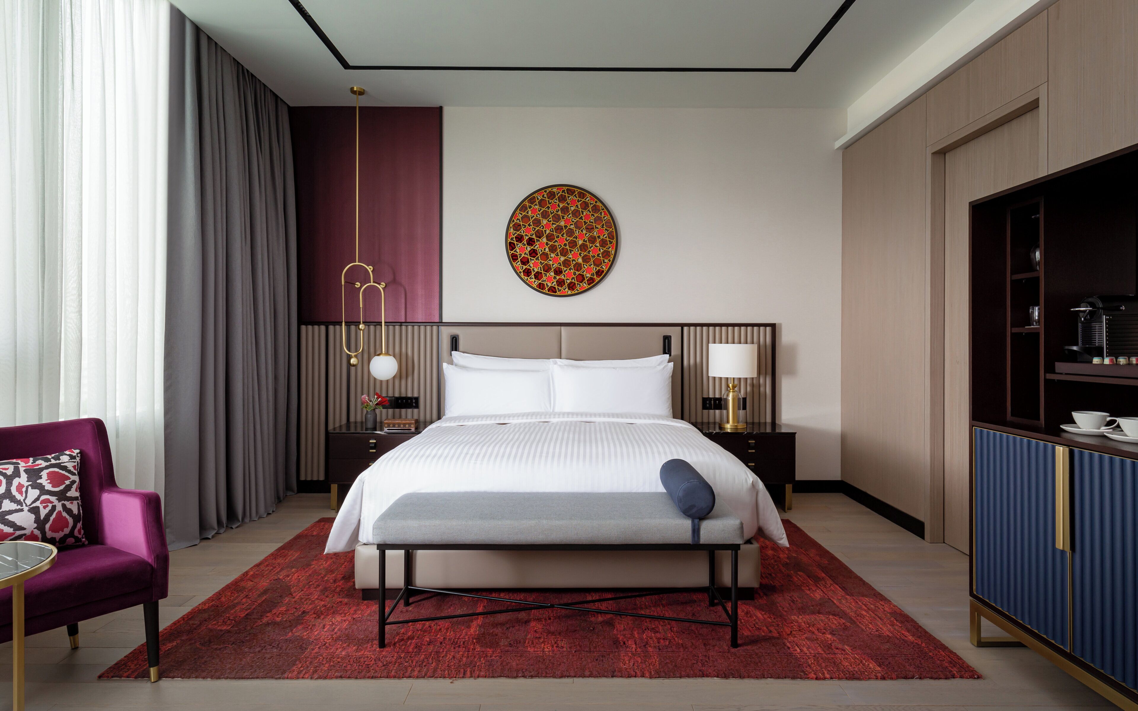 A hotel room with a bed, bedside tables, a chair, a red rug, a wall art piece, and a cabinet.