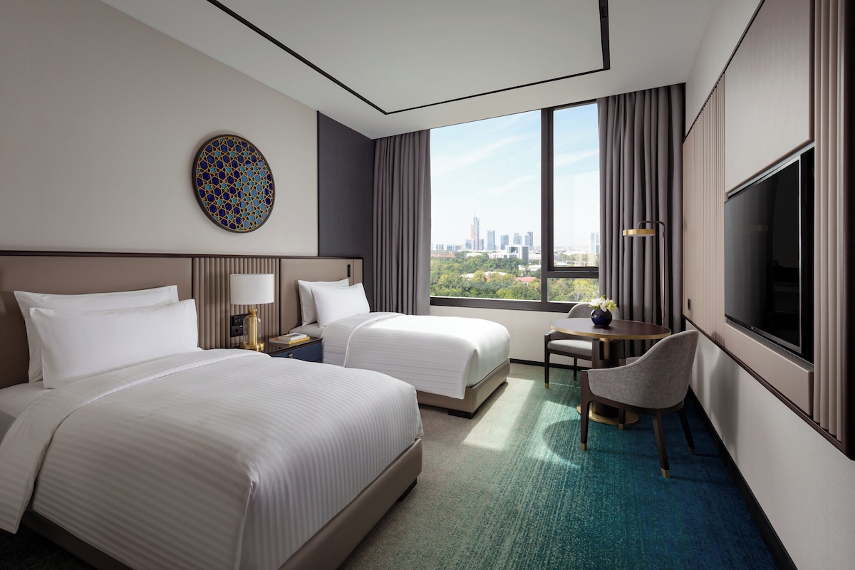 A hotel room with two beds, a desk, a chair, a TV, and a large window with a city view.