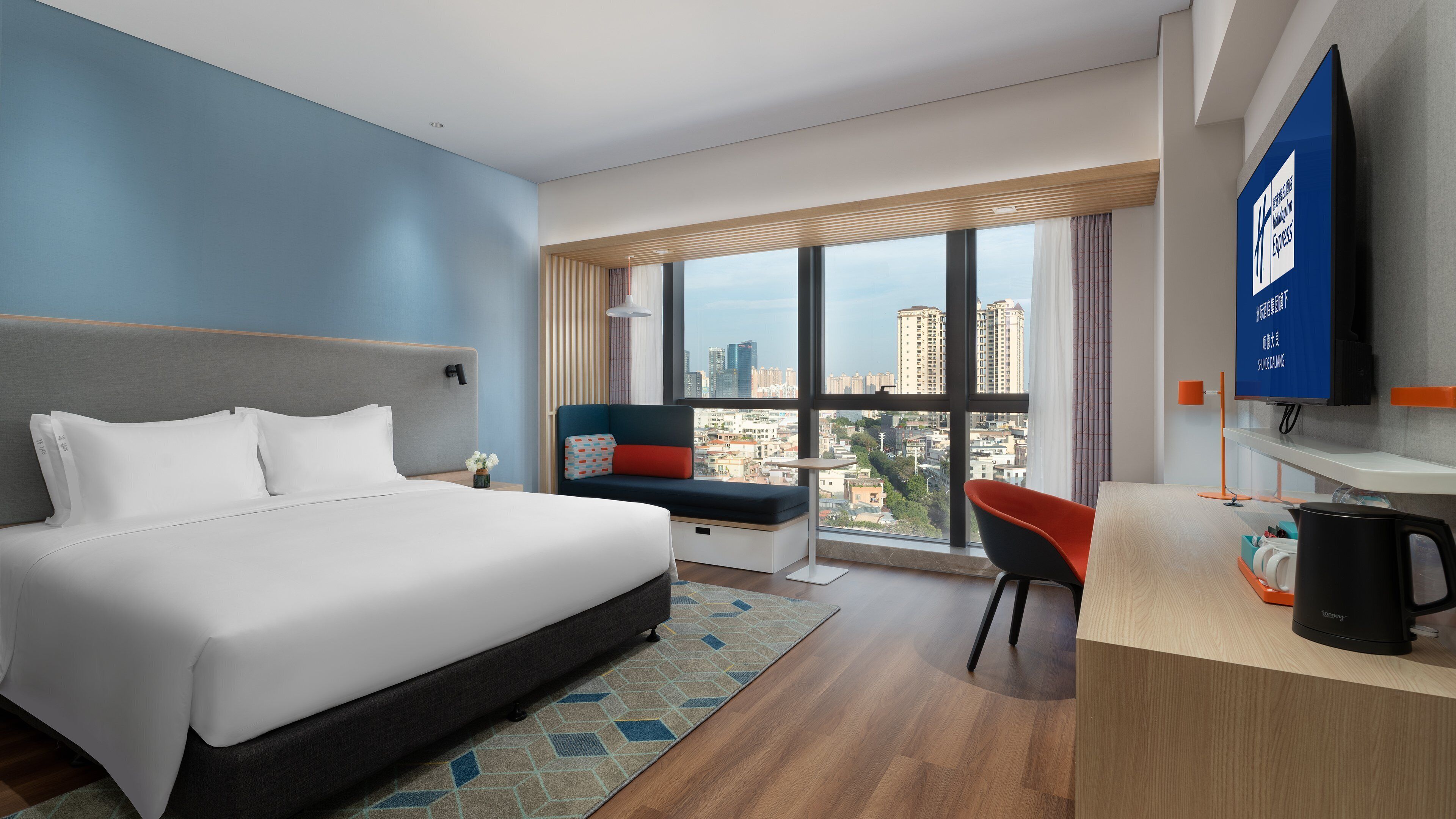 A hotel room with a large bed, a desk with a coffee maker, a view of the city, and a television on the wall.