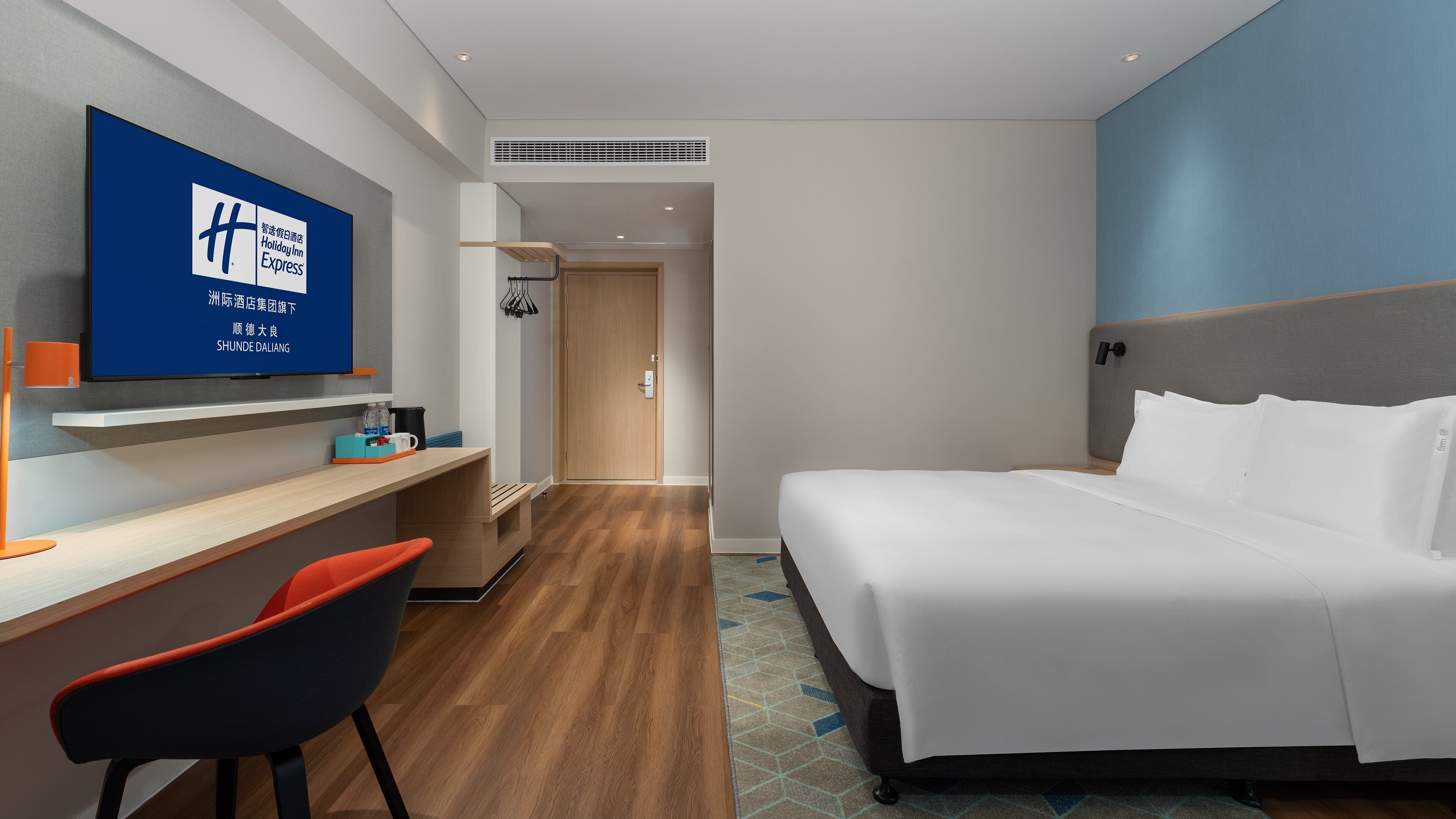 Una habitación de hotel con cama, escritorio con silla, televisor montado en la pared y puerta que da a otra estancia.