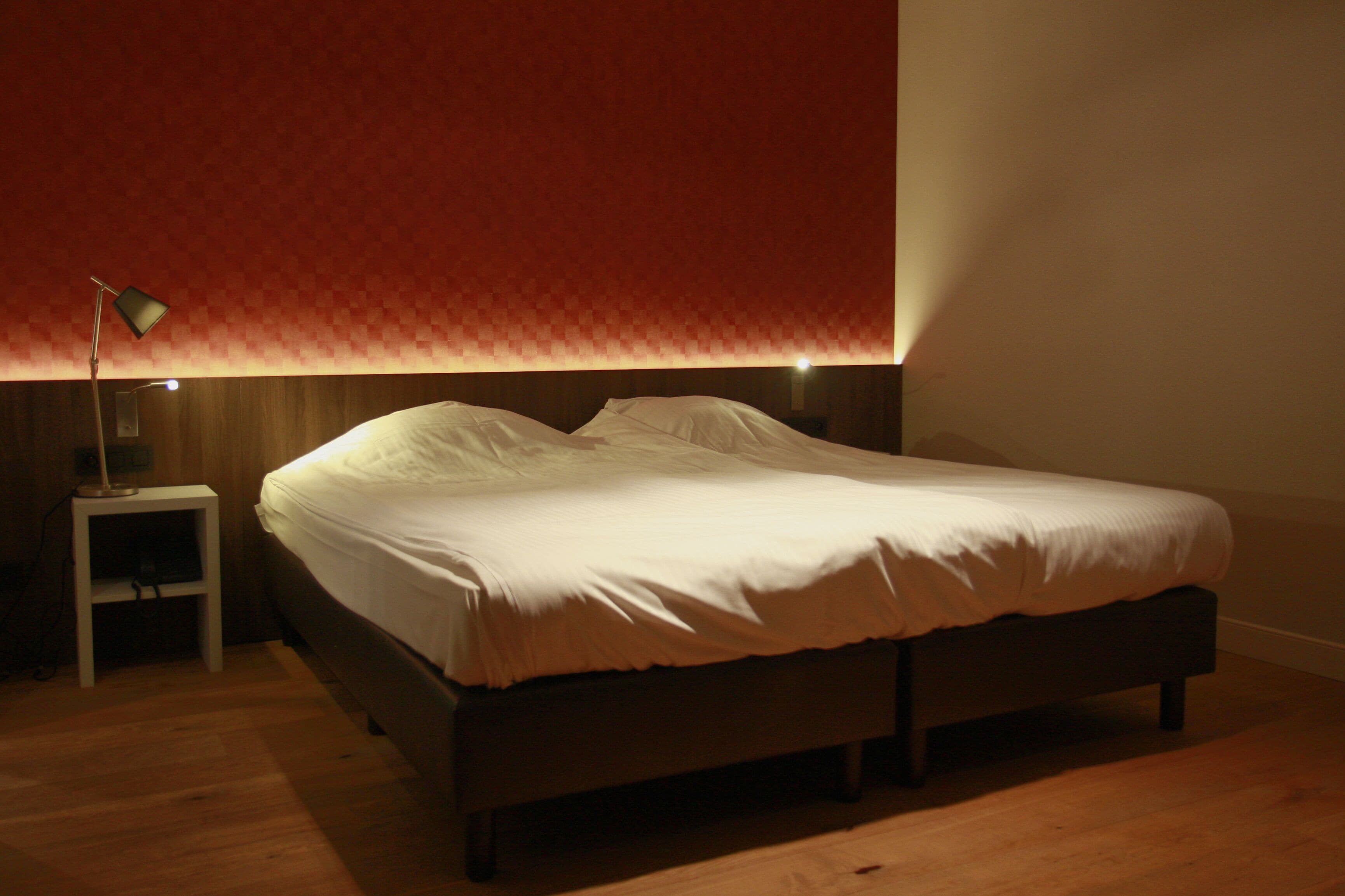 Una cama bien tendida en una habitación con suelo de madera y pared con estampado rojo.