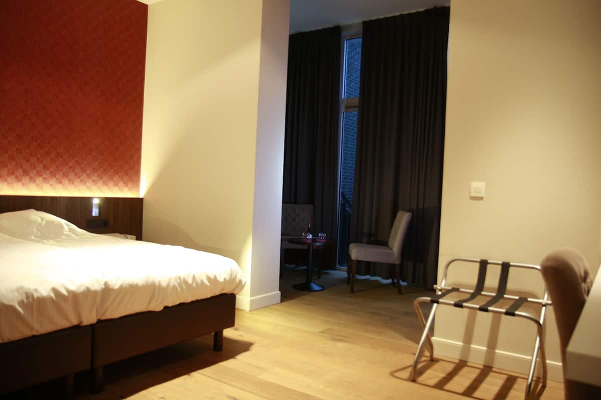 A hotel room with a bed, a chair, a small table, and a window with curtains.