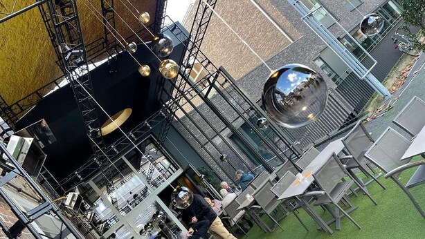 An outdoor dining area with tables and chairs, surrounded by a metal structure and a glass facade. - Hotel de Boskar Pelt (Pelt)