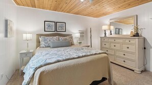 Ein Schlafzimmer mit Holzbalkendecke, einem Bett mit gemusterten Betttextilien, Nachttischlampen, einer Kommode und Wandbildern.