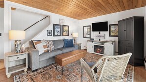 Ein Wohnzimmer mit Sofa, Bank, Korbsessel, Fernseher, Kamin und gerahmten Bildern an der Wand.