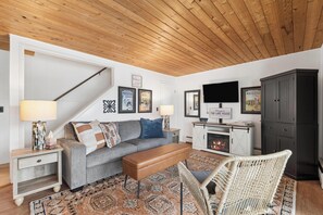 A living room with a sofa, bench, wicker chair, TV, fireplace, and framed pictures on the wall. - Waterfront, Whitefish Lake Condo, 1D Down! (Whitefish)