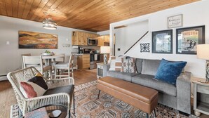Ein modernes Wohnzimmer mit Sofa, Bank und Korbsessel, eine Küche mit Holzausbau und Mikrowelle, sowie ein Essbereich mit Tisch und Stühlen.