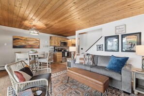 Ein modernes Wohnzimmer mit Sofa, Bank und Korbsessel, eine Küche mit Holzausbau und Mikrowelle, sowie ein Essbereich mit Tisch und Stühlen.