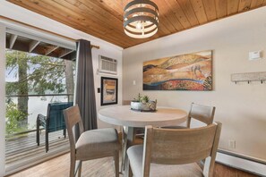 A dining area with a round table, chairs, a pendant light, a painting, and a view of trees through a window. - Waterfront, Whitefish Lake Condo, 1D Down! (Whitefish)