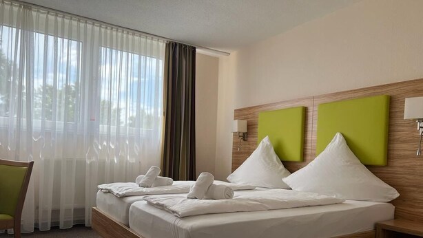 Ein Hotelzimmer mit einem großen Bett, einem hölzernen Kopfteil, einer grunen Akzentwand und einem Fenster mit transparenten Vorhängen.