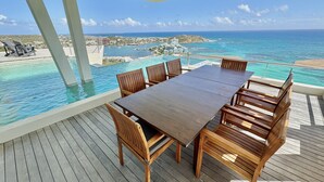 Eine Holzterrasse mit Tisch und Stühlen mit Blick auf einen Pool und den Ozean.
