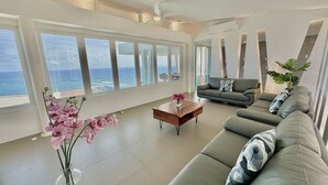 Ein geräumiges Wohnzimmer mit großem Fenster mit Meerblick, einem grauen Sofa, einem Holztisch und einer Vase mit rosa Orchideen.