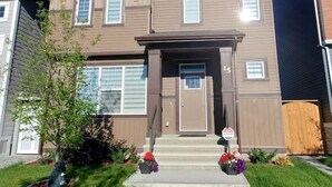 A two-story residential building with a brown exterior, a front porch, and a small garden with flowers. - Delightful 1-Bedroom Private Suite in a New Home (Calgary)