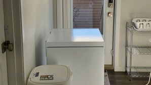 A laundry room with a washing machine, a trash bin, and a metal shelf unit. - Delightful 1-Bedroom Private Suite in a New Home (Calgary)