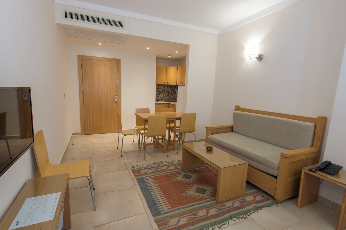 Un espacio habitable compacto con sofá, mesa de comedor con sillas, una pequeña mesa de centro, televisor y una zona de cocina al fondo.