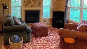 A cozy cabin interior with wooden walls, a fireplace, two sofas, a TV, and large windows offering a view of the outdoors.