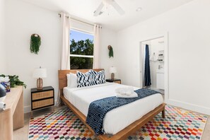 Ein modernes Schlafzimmer mit einem hölzernen Bett, weißer Bettwäsche, einem bunten Karotten Teppich und einem Fenster mit Vorhängen.