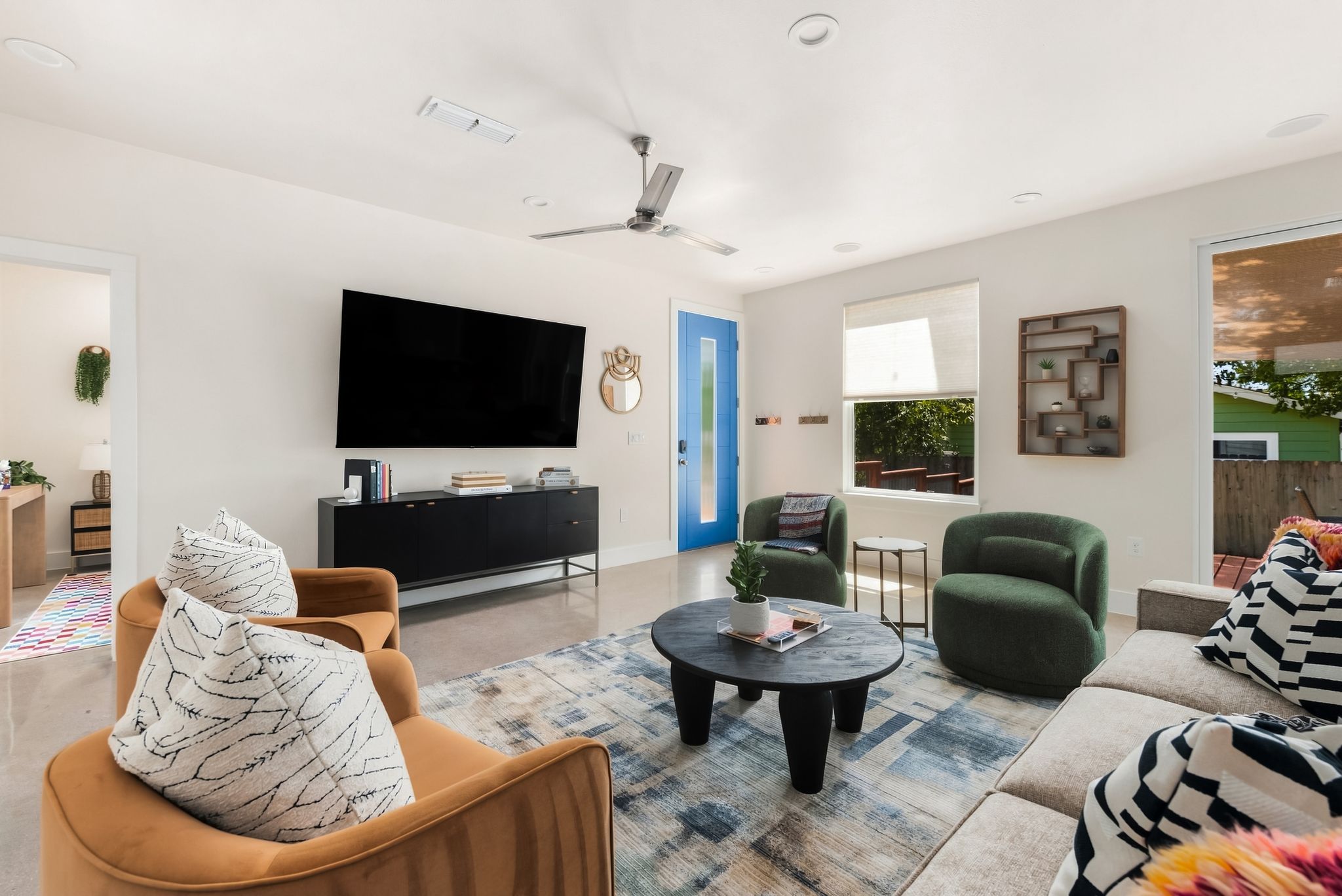 A modern living room with a flat-screen TV, a ceiling fan, a blue door, and a view of the outdoors.