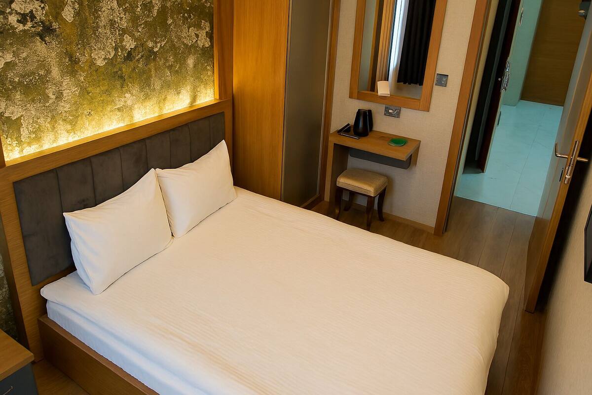 A hotel room with a bed, a small stool, a nightstand, a mirror, and a wall with a textured surface.