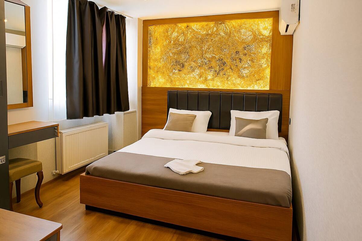 A hotel room with a large bed, a wooden headboard, a bedside table, a mirror, and a window with curtains.