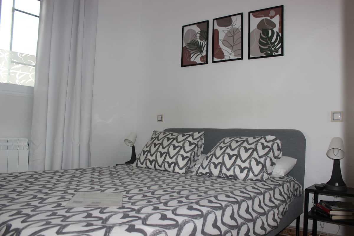 A bedroom with a bed, heart-patterned pillows, a bedside table with a lamp, and three framed artworks on the wall.