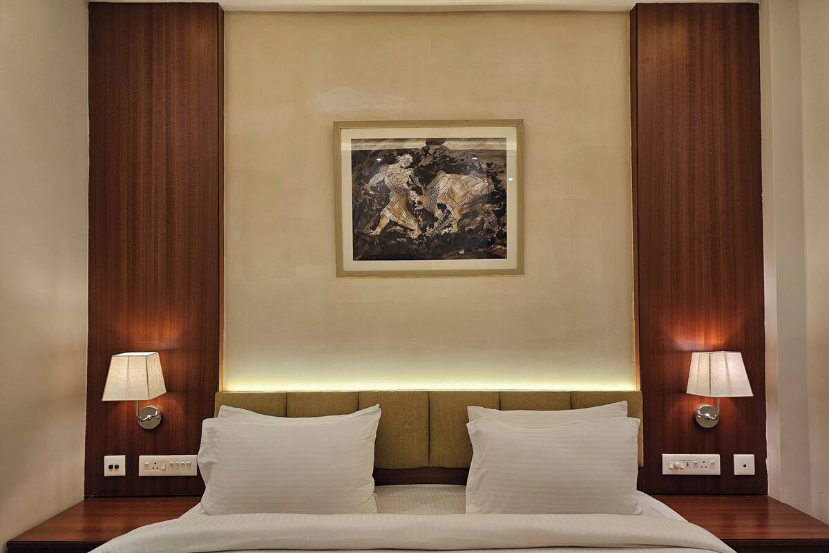 A hotel room with a bed, two bedside lamps, a wall-mounted painting, and a wooden headboard.
