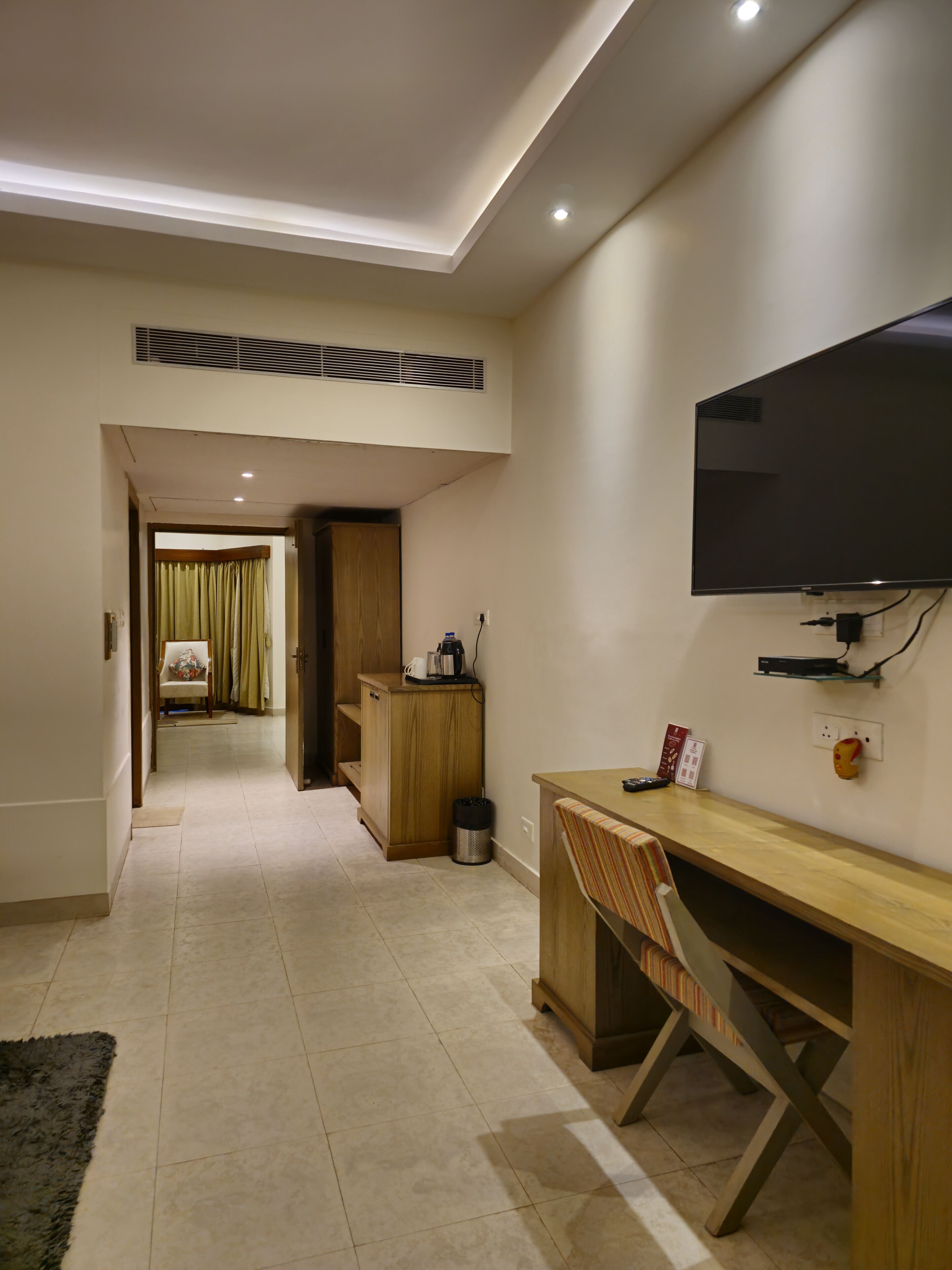 Habitación de hotel moderna con un escritorio de madera, un televisor montado en la pared, una kitchenette pequeña y vistas al pasillo.