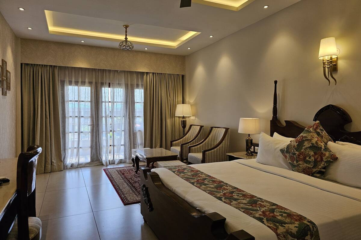 A hotel room with a large bed, two bedside tables with lamps, a ceiling fan, and a view of the outdoors through large windows.