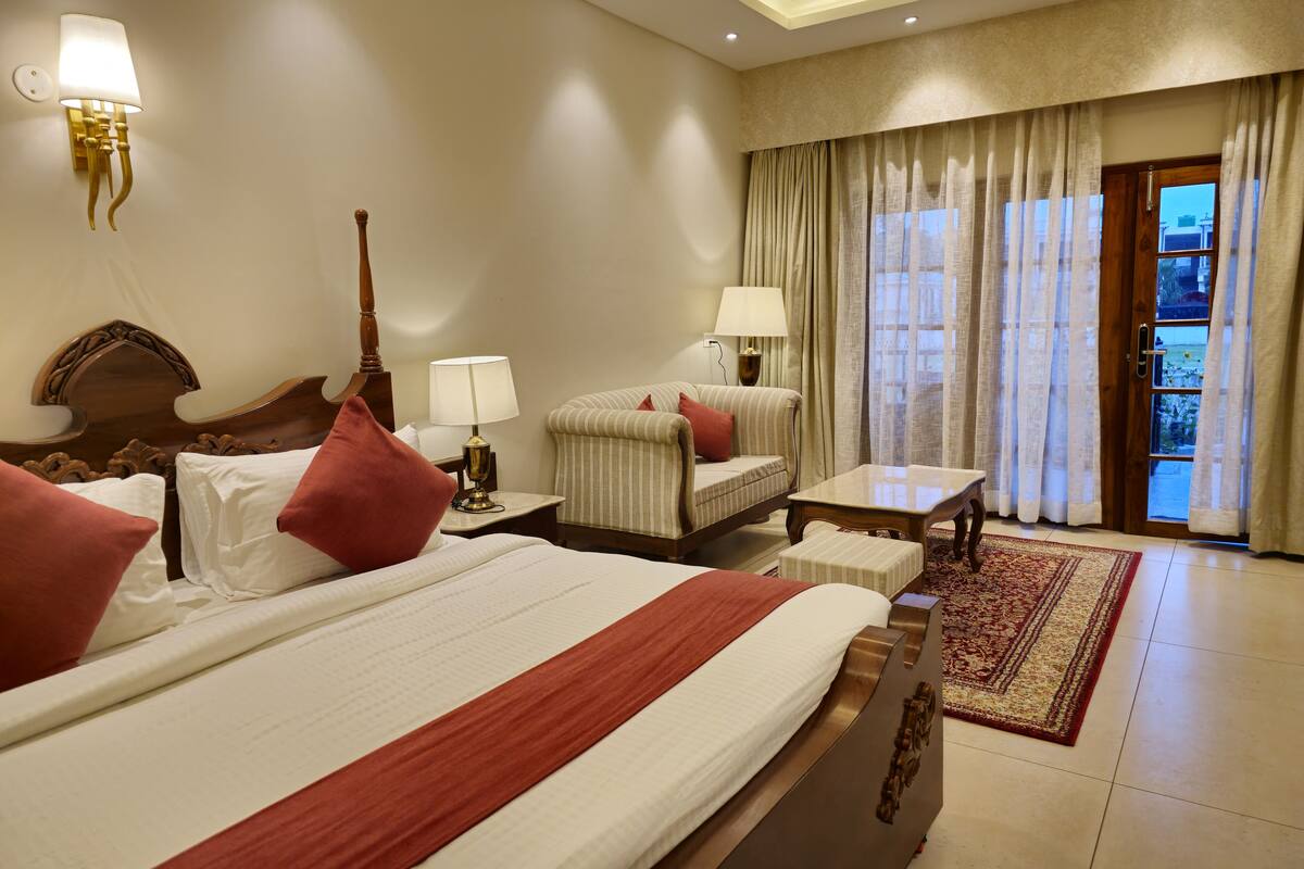 A hotel room with a large bed, two bedside tables, a sofa, a small table, and a view of the outdoors through a glass door.