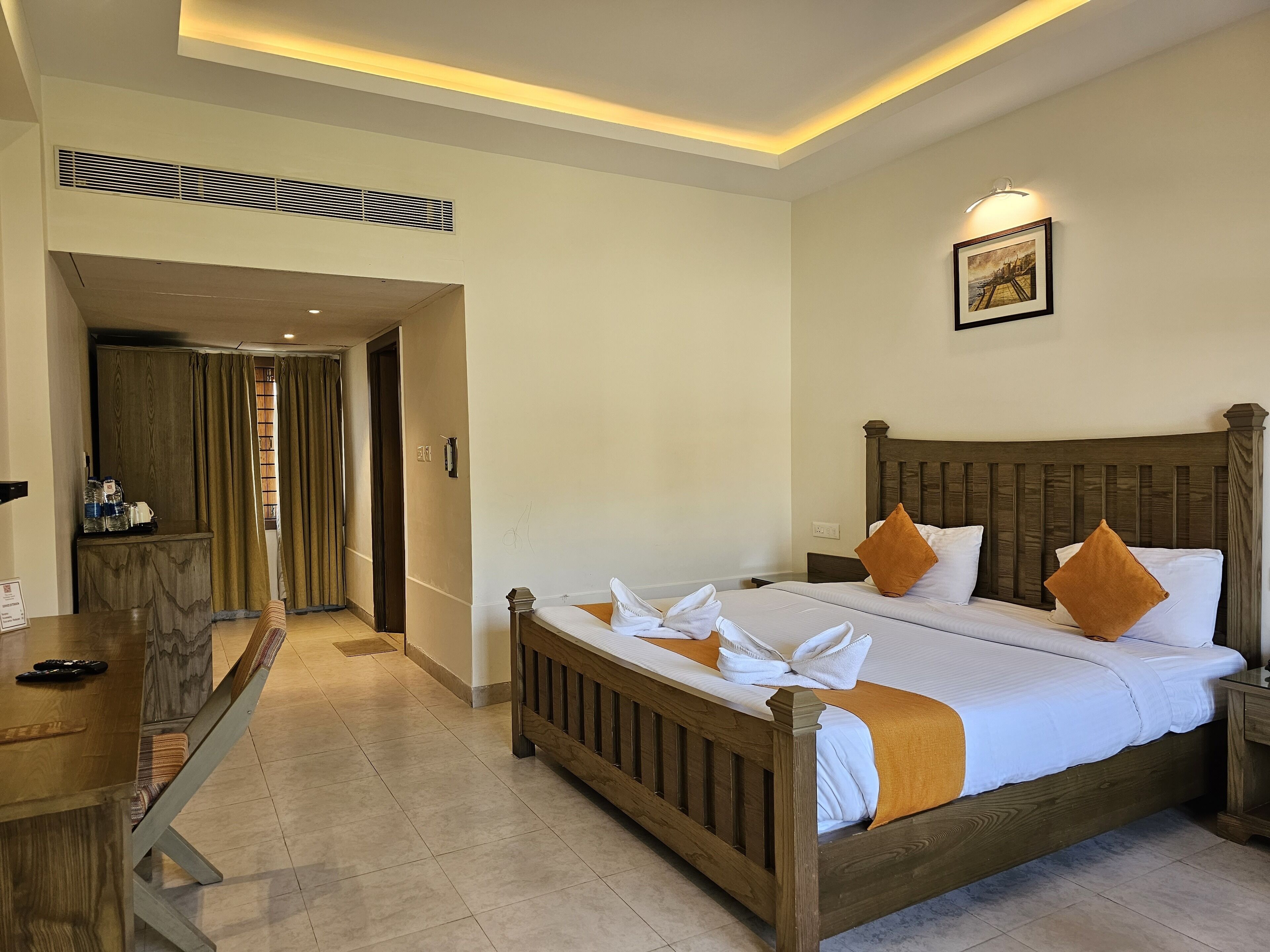 A hotel room with a wooden bed, a desk with a chair, a bathroom with a shower, and a hallway with a door.