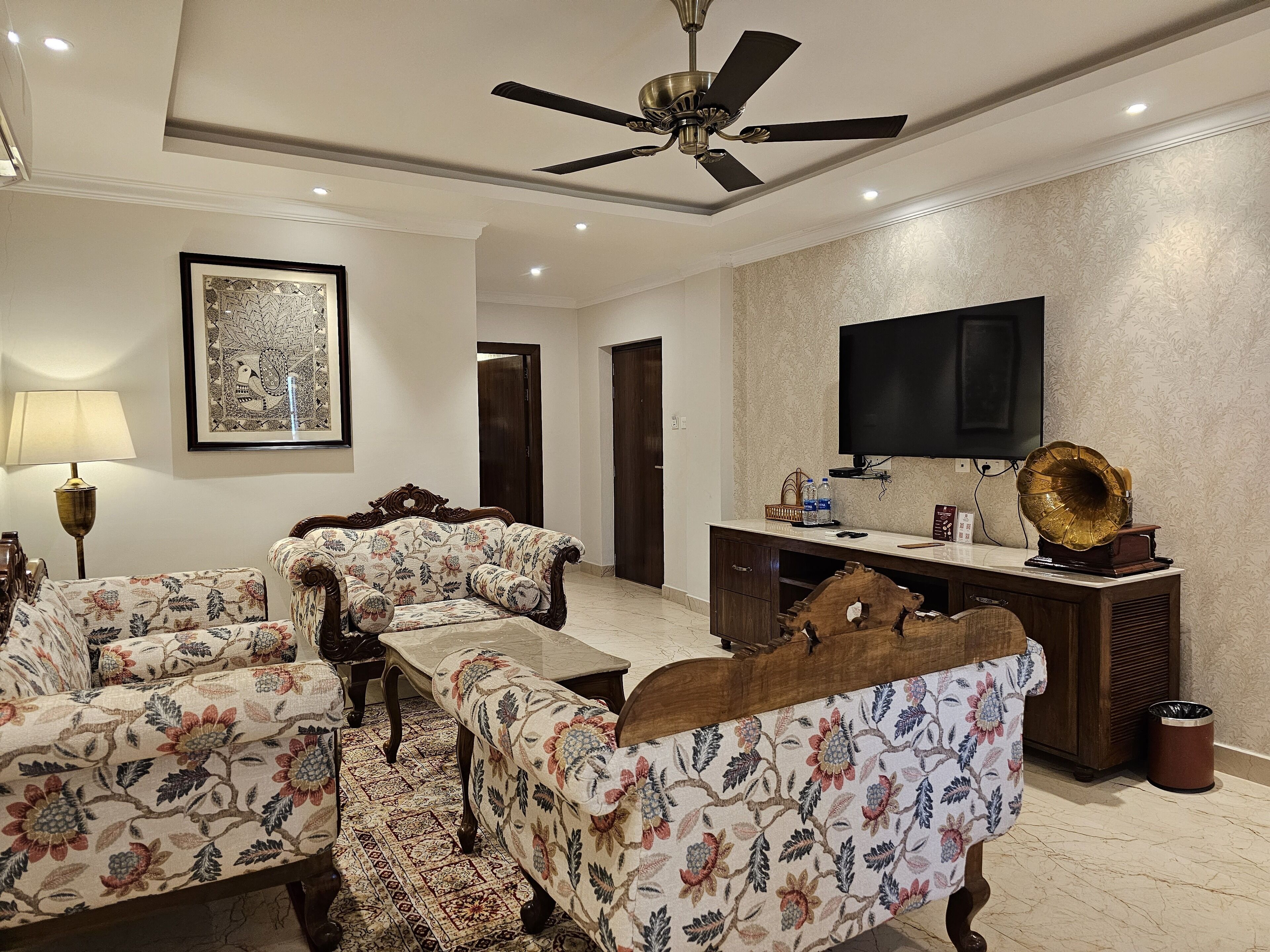 A spacious living room with a ceiling fan, a large flat-screen TV, a vintage gramophone, and a floral-patterned sofa set.