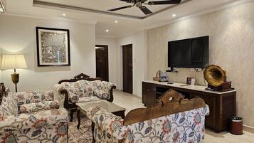 A spacious living room with a ceiling fan, a large flat-screen TV, a vintage gramophone, and a floral-patterned sofa set.