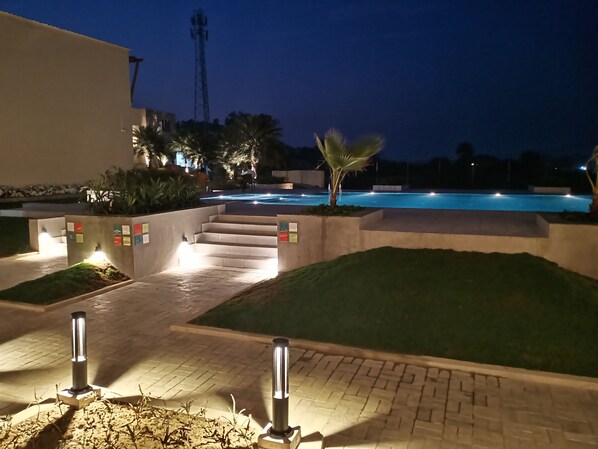 Vista nocturna de una zona de piscina con escalones de acceso, rodeada de vegetación e iluminada por luces de camino.