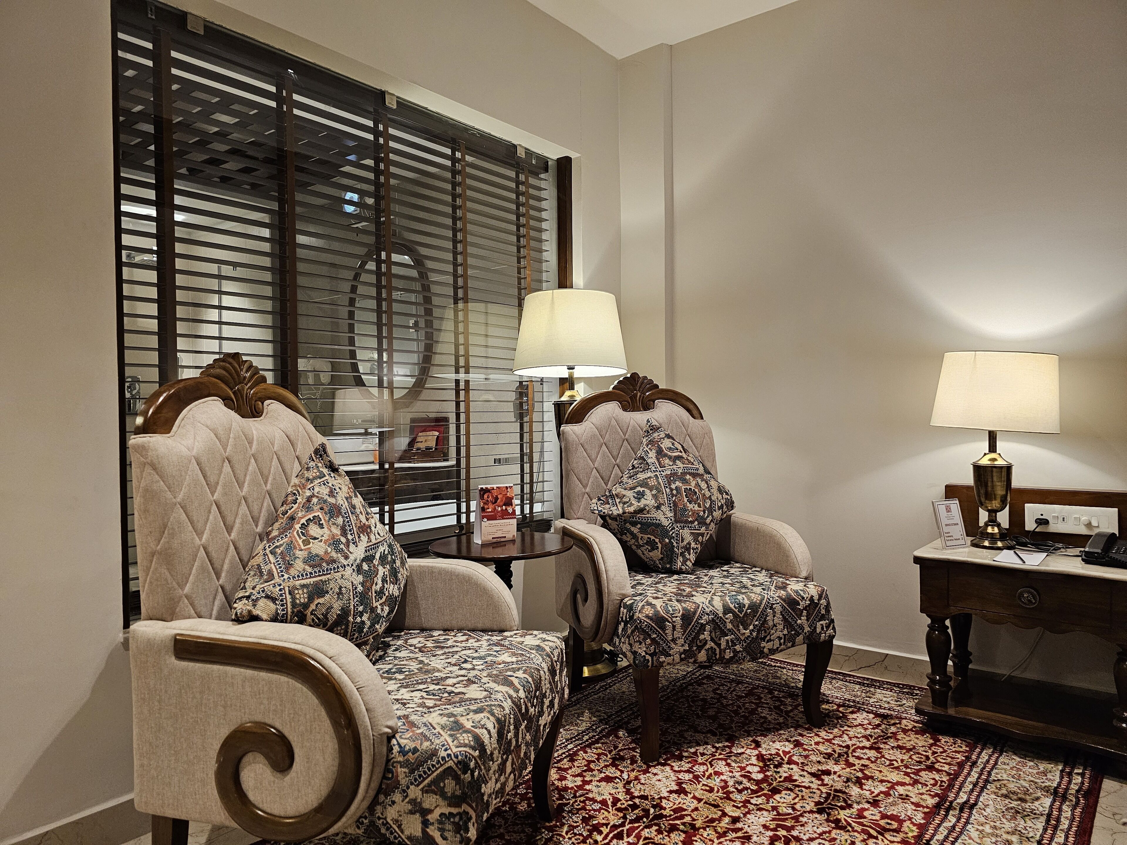 Een hotelkamer met twee gestoffeerde fauteuils, een bijzettafeltje met een lamp en een groot raam met jaloezieën.