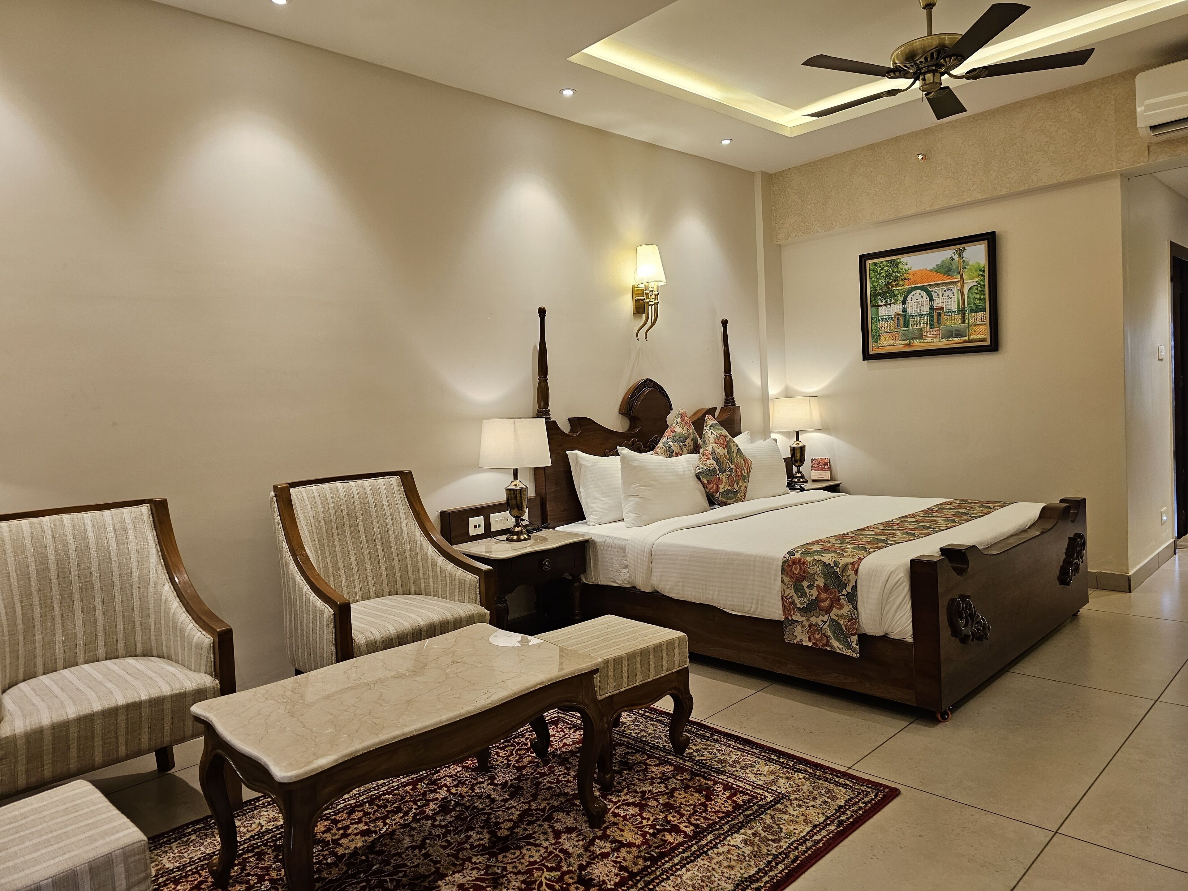 A hotel room with a large bed, two bedside tables with lamps, a ceiling fan, a painting on the wall, and a patterned rug.