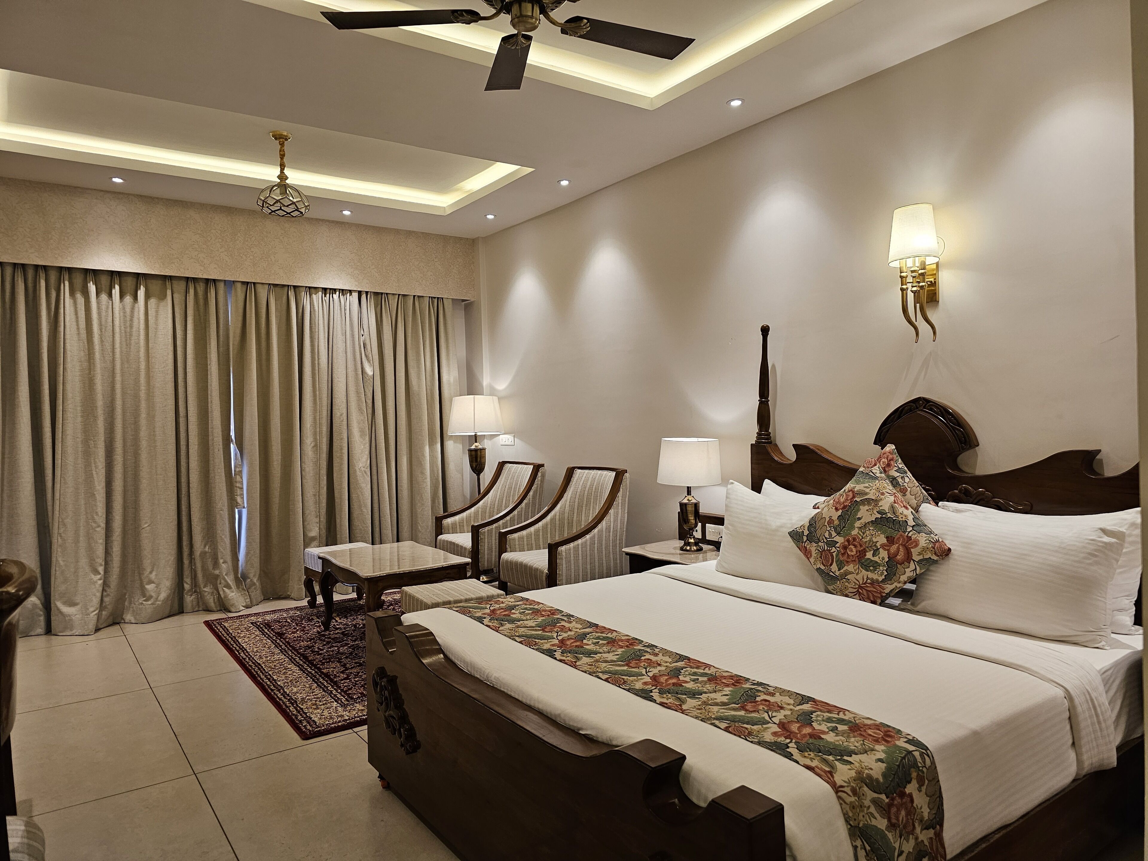 A hotel room with a large bed, two armchairs, a small table, a ceiling fan, and a nightstand with lamps.