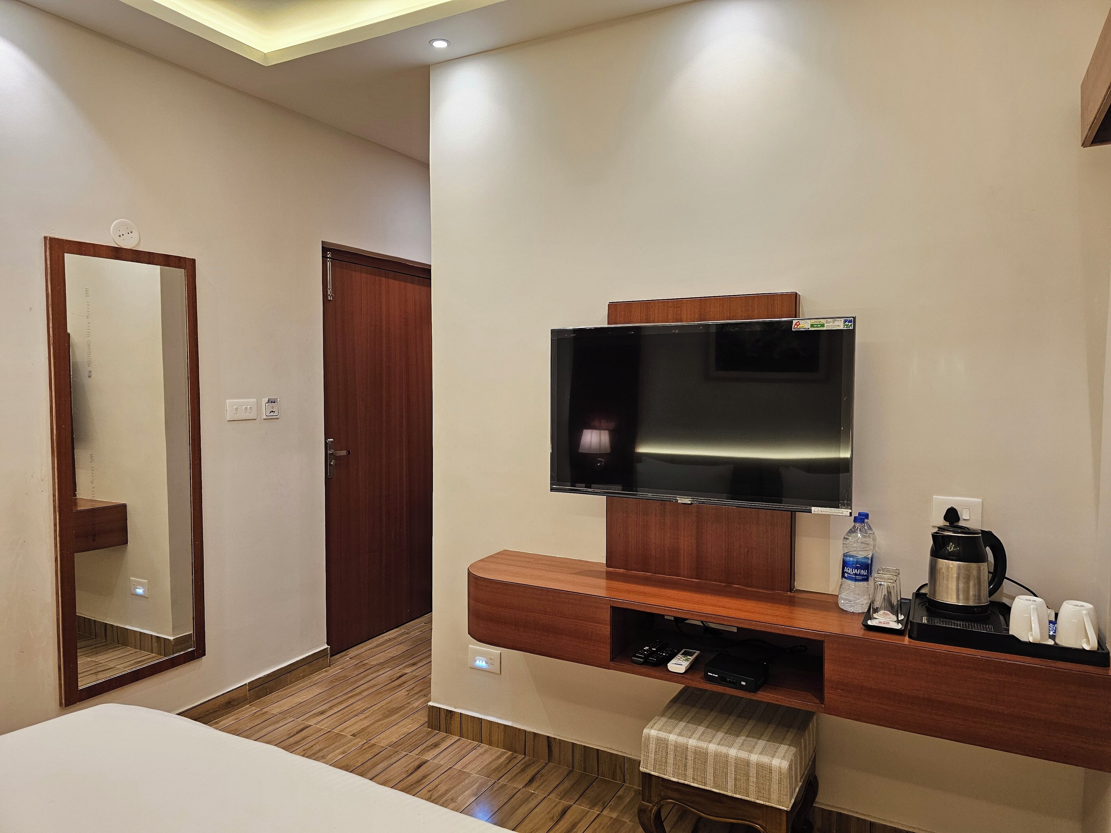 A hotel room with a wooden floor, a flat-screen TV mounted on a wooden cabinet, a small table with a checkered tablecloth, and a coffee maker.