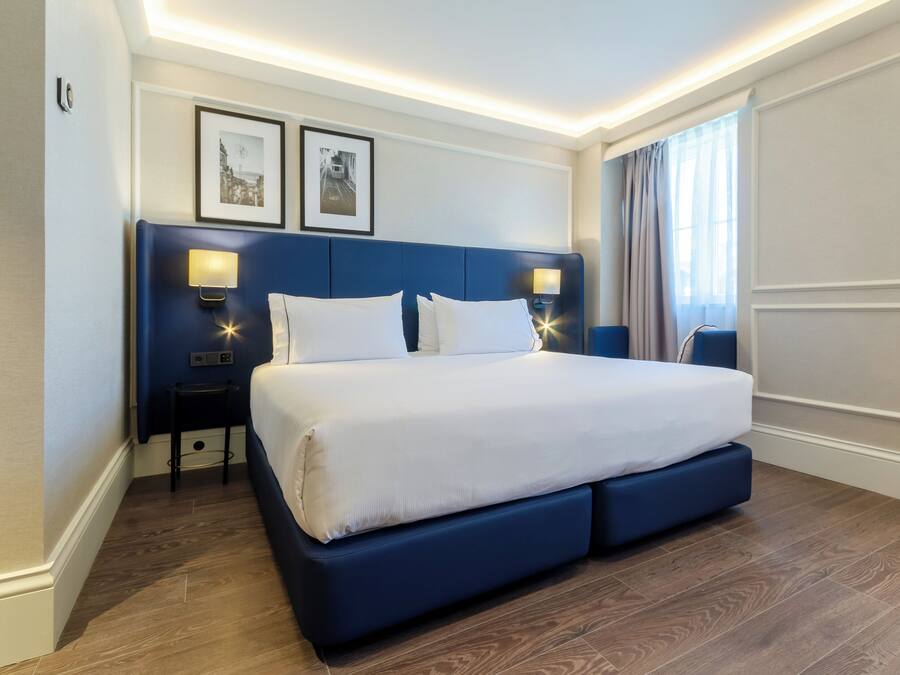 Una habitación de hotel moderna con una cama grande, dos mesitas de noche con lámparas, un cabecero y cuadros enmarcados en la pared.