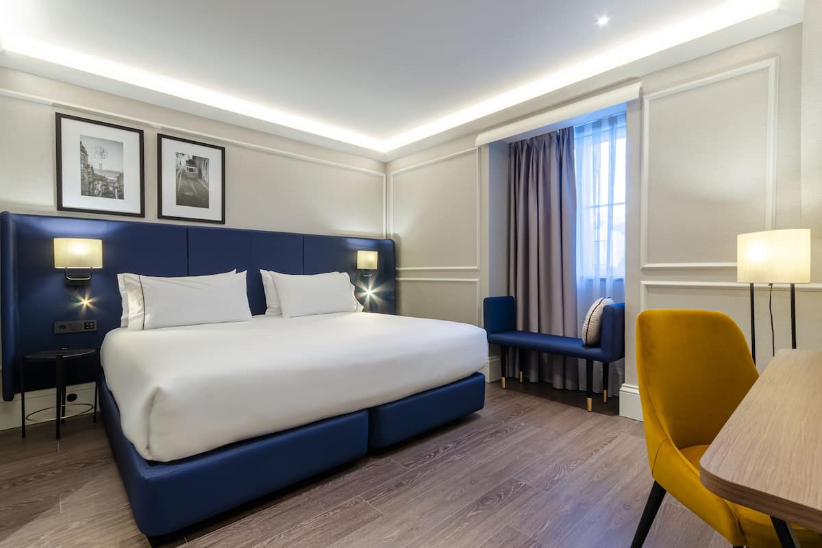 Habitación de hotel con cama, mesitas de noche, un escritorio, una silla y una ventana con cortinas.