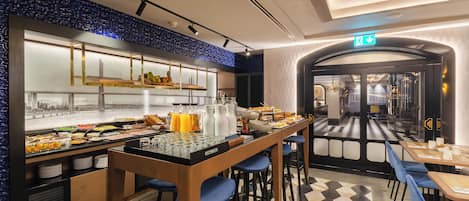 A breakfast bar with various food items and beverages, a seating area with blue chairs, and a checkered floor pattern.