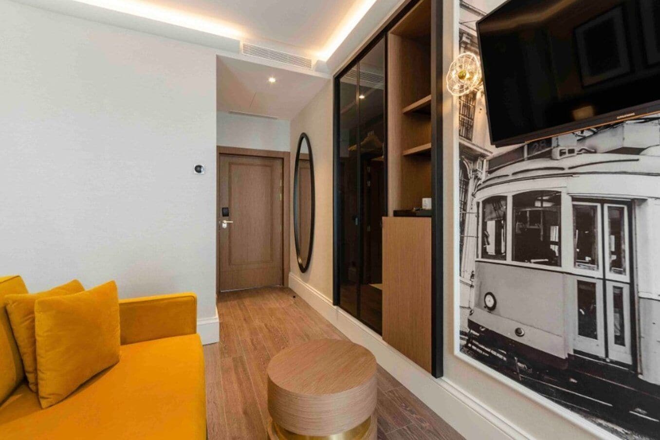 Una habitación de hotel moderna con un sofá amarillo, un taburete de madera redondo, un espejo grande y un televisor instalado en la pared.