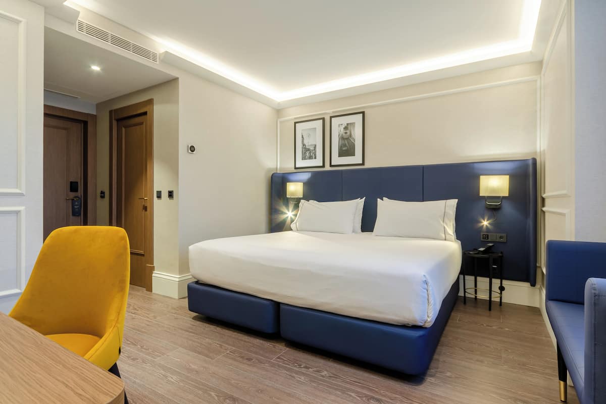 Una habitación de hotel con una cama grande, una silla amarilla, una mesita y dos lámparas de pared.