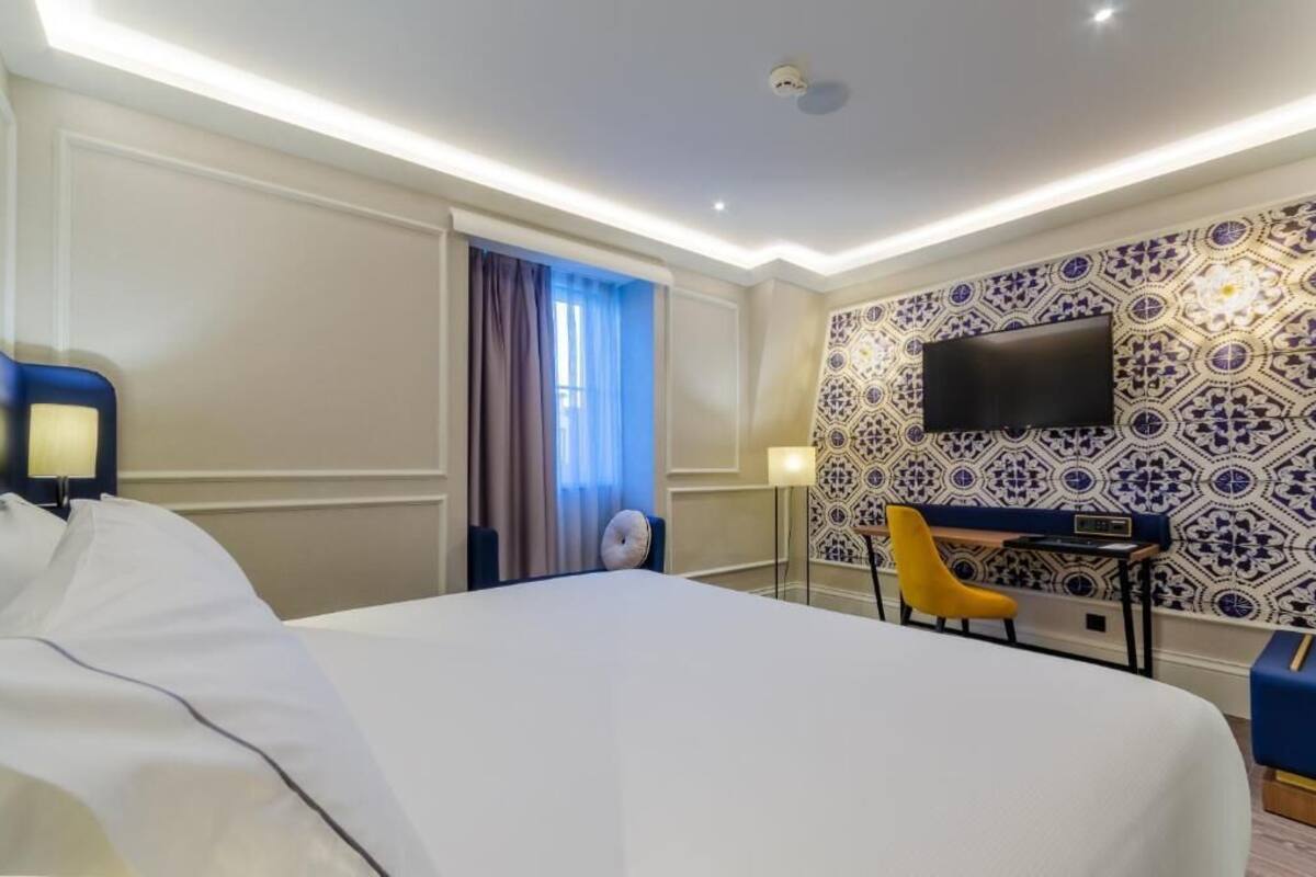 Habitación de hotel con una cama grande, un escritorio con una silla, un televisor de pantalla plana montado en la pared y un diseño de baldosas con motivos en la pared.