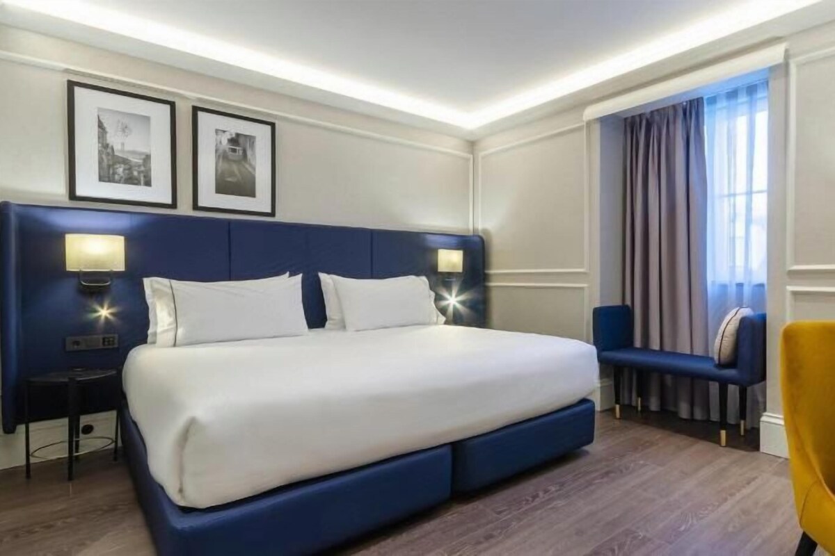 Een hotelkamer met een groot bed, nachtlampjes, een bankje, een stoel en een raam met gordijnen.